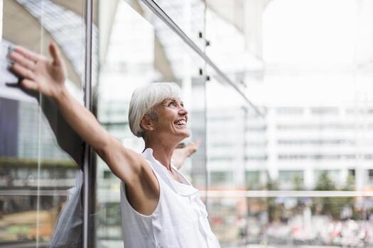 Happy senior woman leaning against glass facade with outstretched arms photo
