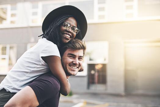 Young playful interracial couple playing, bonding and hugging while standing in the city together on weekend. Happy husband giving wife a piggyback ride, enjoying a day in the urban town or traveling photo